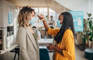 Two women face each other in an office. with protective face masks using infrared thermometer for measuring temperature before entrance in office. Women on left scans other with forehead thermometer.